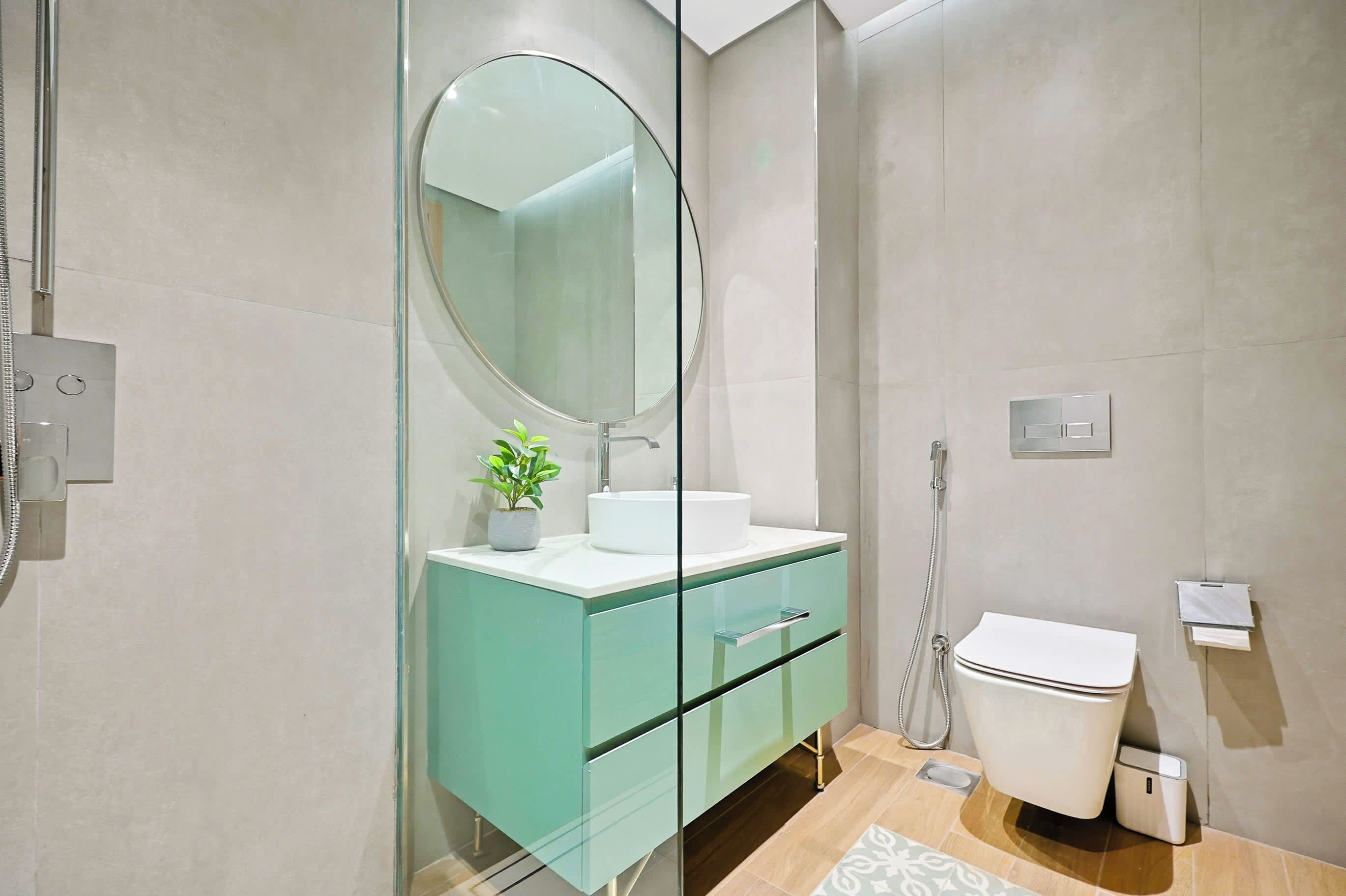 Et moderne badeværelse med et glasbadeværelsesafskærmning, et hvidt toilet og en håndvask med et rundt spejl og en potteplante.