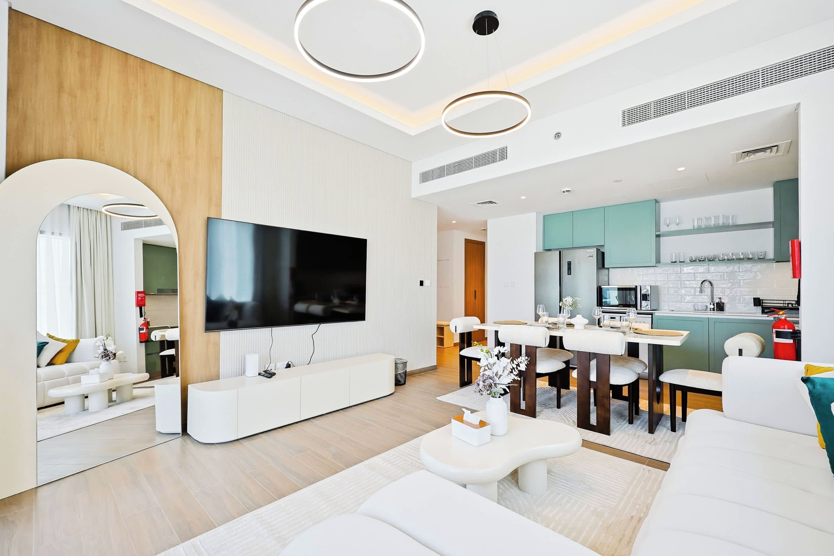 A modern living room with a flat-screen TV, white furniture, and a kitchen area with teal cabinets.