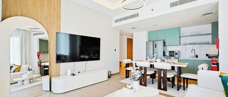 Sala moderna com televisão de tela plana, móveis brancos e área de cozinha com armários verde-água.