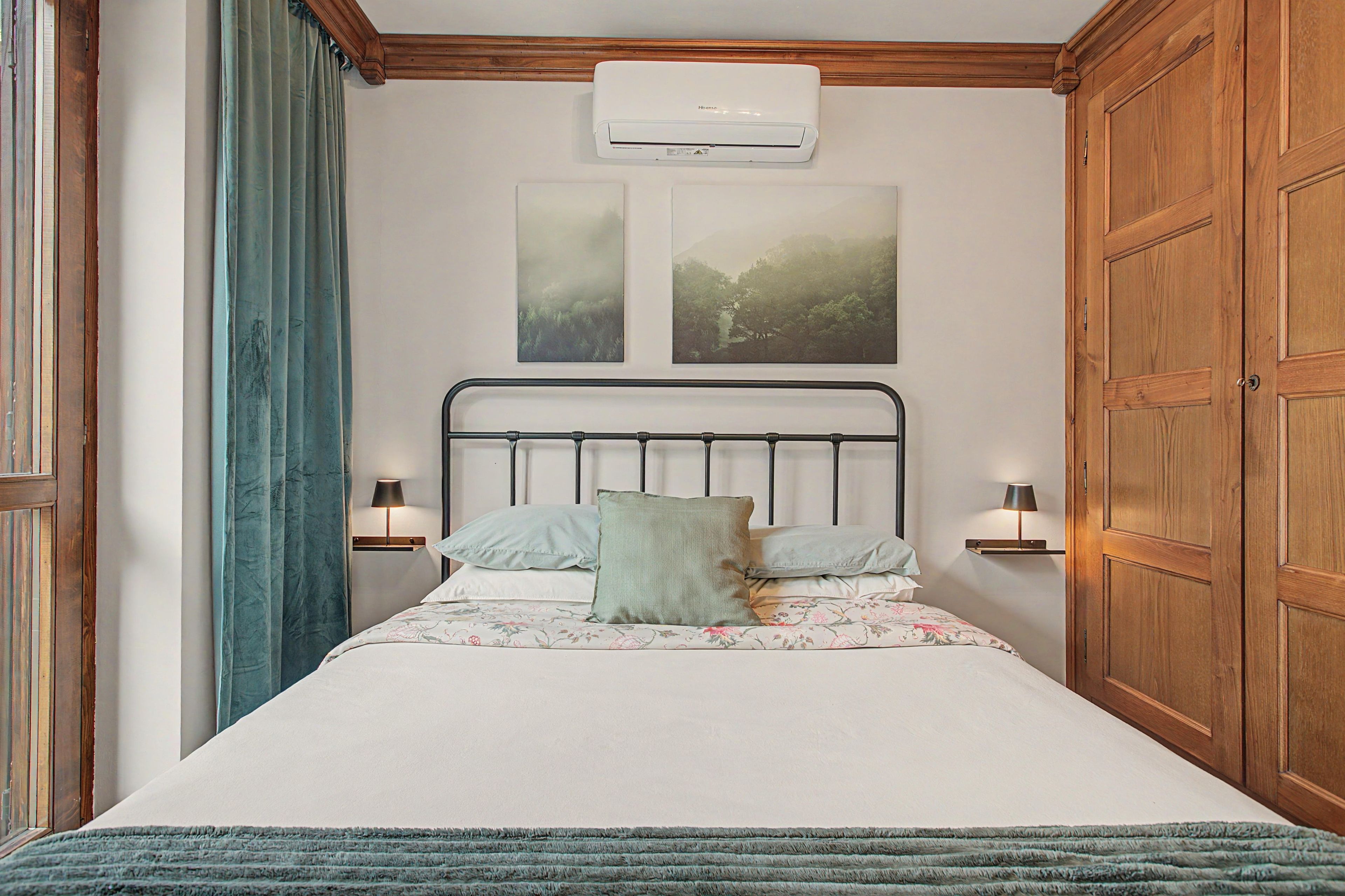 A bedroom with a bed, two wall-mounted lamps, a wooden wardrobe, and two framed pictures.