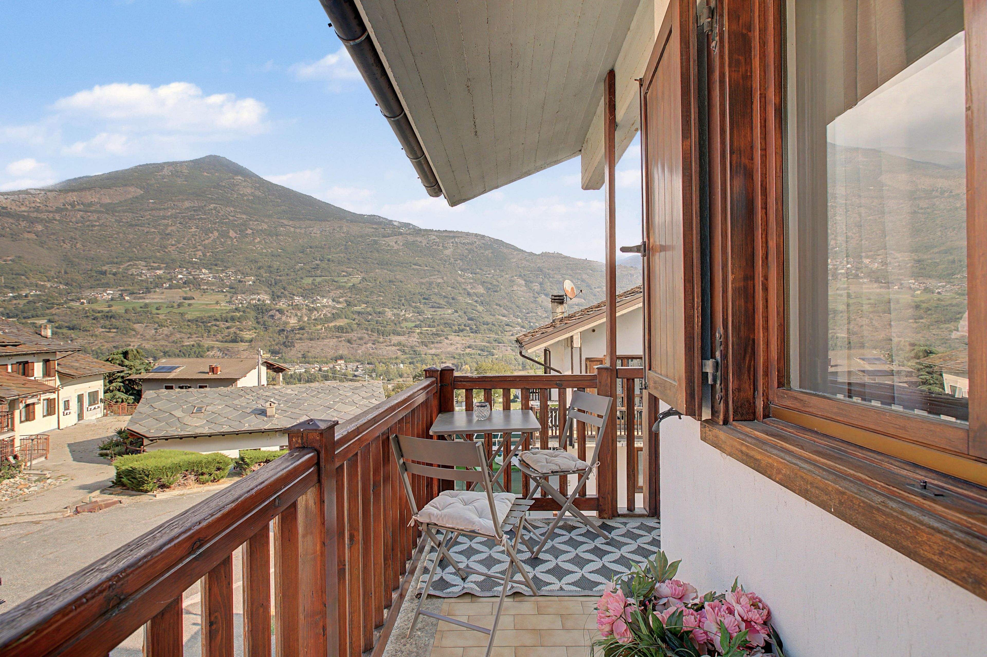Ein Balkon mit Tisch und Stühlen, mit Blick auf eine Berglandschaft und Häuser im Tal.