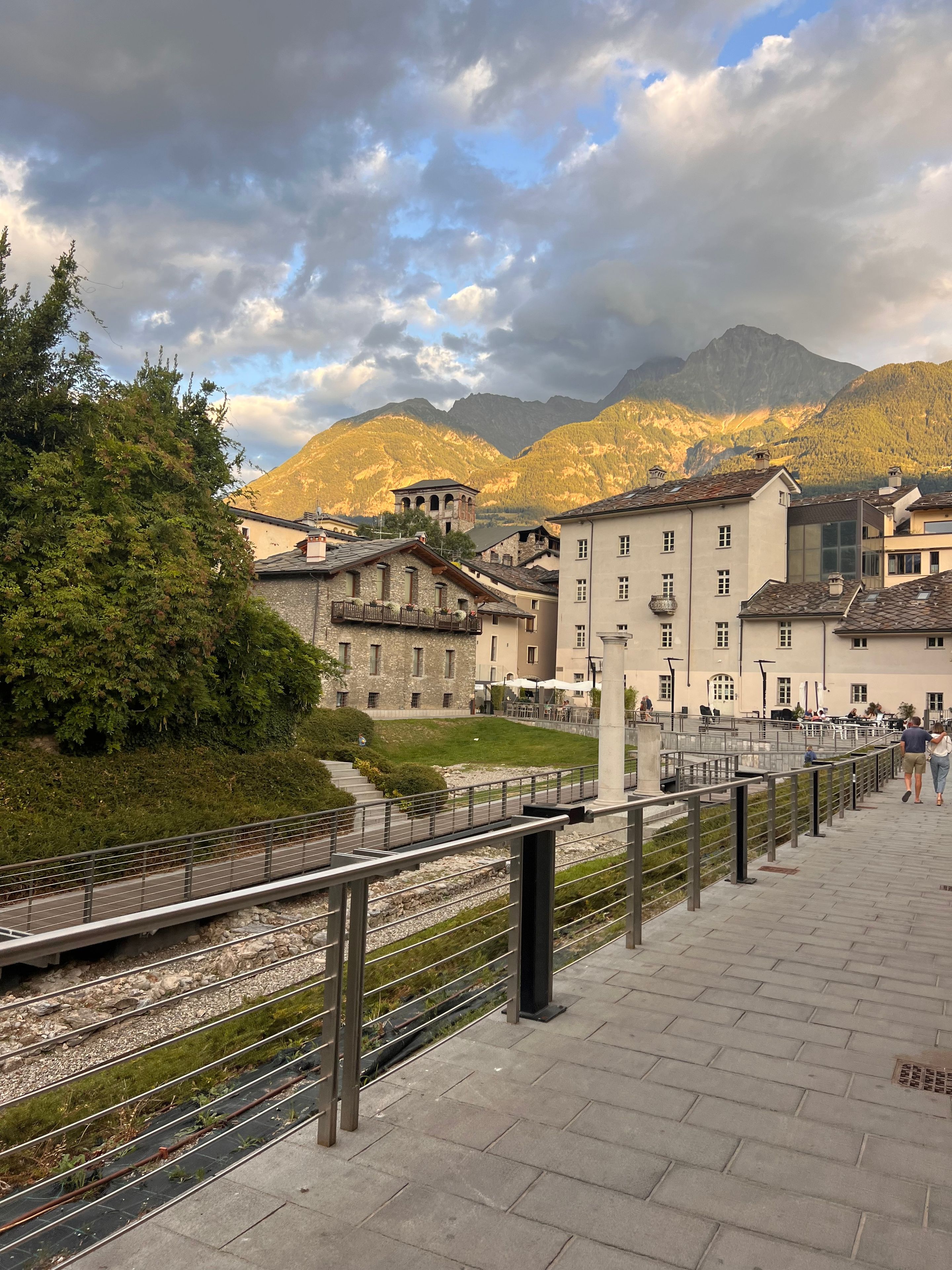 Un percorso pedonale con vialetto in pietra, ringhiera metallica e vista su edifici tradizionali con montagne sullo sfondo.