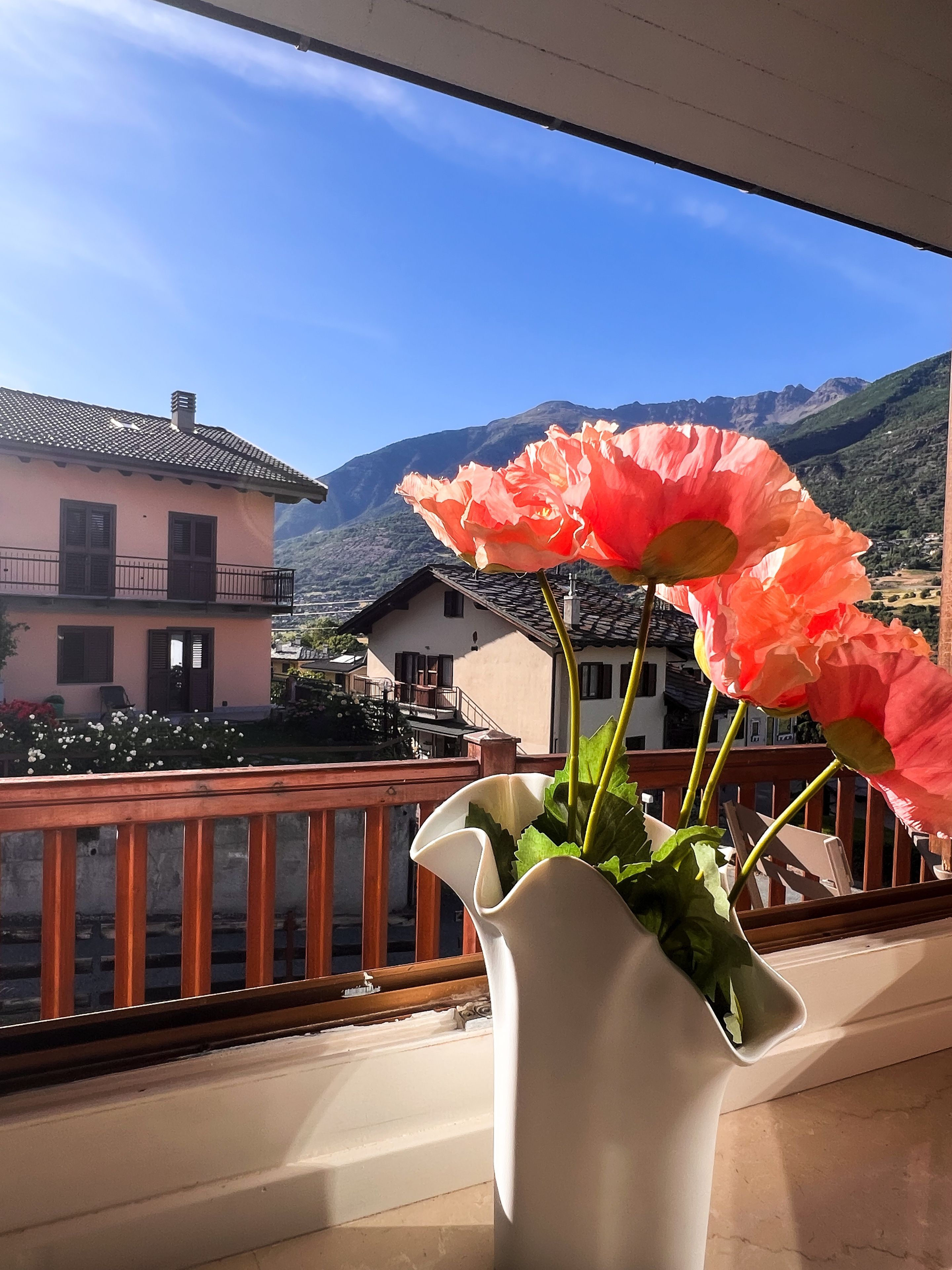 A balcony with a vase of flowers, a railing, and a view of houses and mountains.