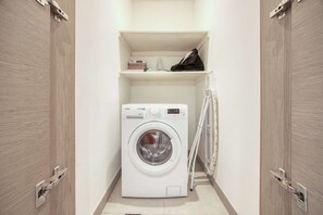 Ein kleiner Hauswirtschaftsraum mit einer Waschmaschine mit Frontlader, einem Regal darüber und einem Wäscheständer zur Rechten.