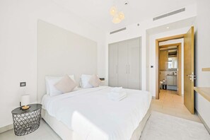 A modern bedroom with a large bed, bedside tables, a wardrobe, and a bathroom visible through an open door. - Sobha Waves 1BR · Pool & Gym · MBR City (Dubai)