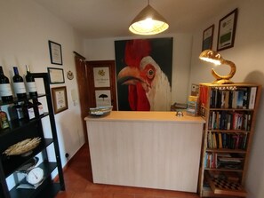 Een receptiebalie met een groot schilderij van een haan, een wijnflespresentatie en een boekenplank.