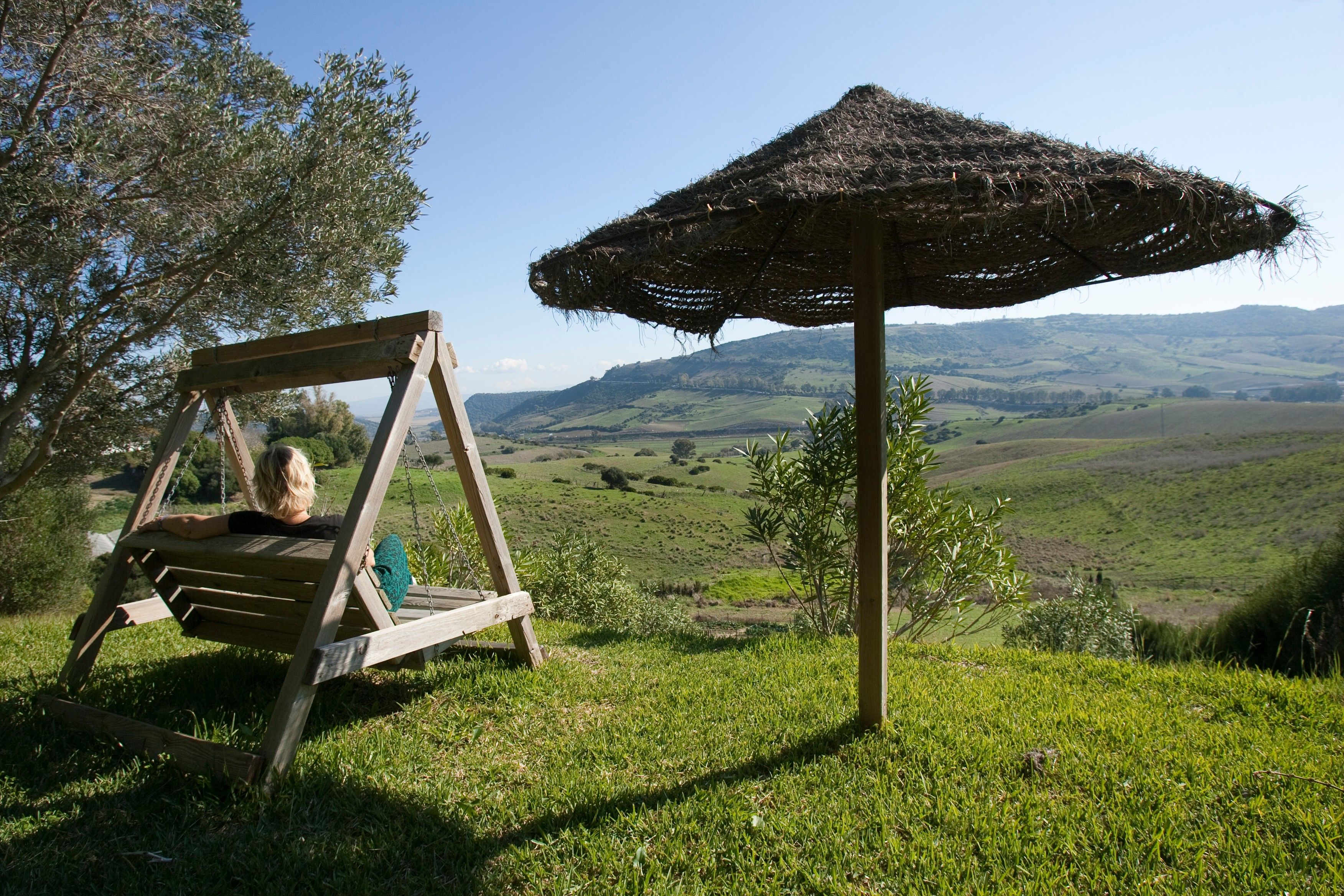 Una silla mecedora de madera y un parasol de paja en una colina cubierta de césped, con vistas a colinas ondulantes al fondo.