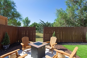 A backyard with a wooden deck, two Adirondack chairs, a square table, and a small fire pit. - Cozy Duplex by Portal Park | Pet Friendly (Colorado Springs)