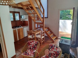 Eine kompakte Küche mit Mikrowelle, Herd und kleinem Kühlschrank. Eine Holztreppe führt zu einer oberen Ebene. Zwei Korbstühle mit Blumenkissen stehen im Wohnbereich.