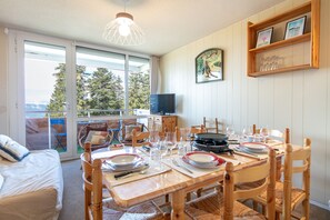 A dining area with a table set for four, a television on a stand, and a view of the outdoors through large windows. - Le Sunset - station de ski accessible à pied (Chamrousse)