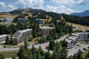 A multi-story hotel with a parking lot, surrounded by trees and mountains in the background. - Le Sunset - station de ski accessible à pied (Chamrousse)