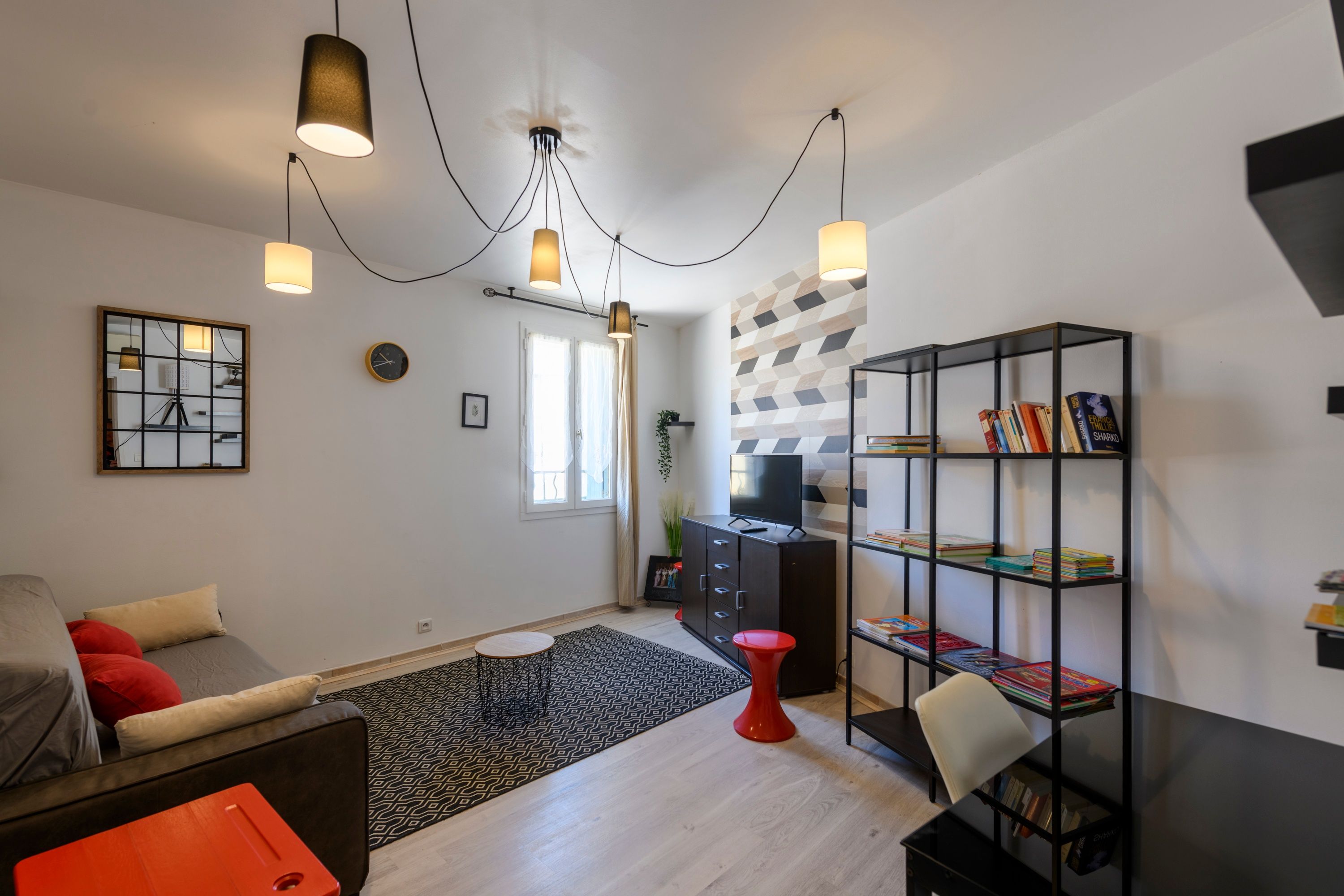 Una sala de estar moderna con un sofá, un taburete rojo, una estantería y un televisor sobre un mueble.