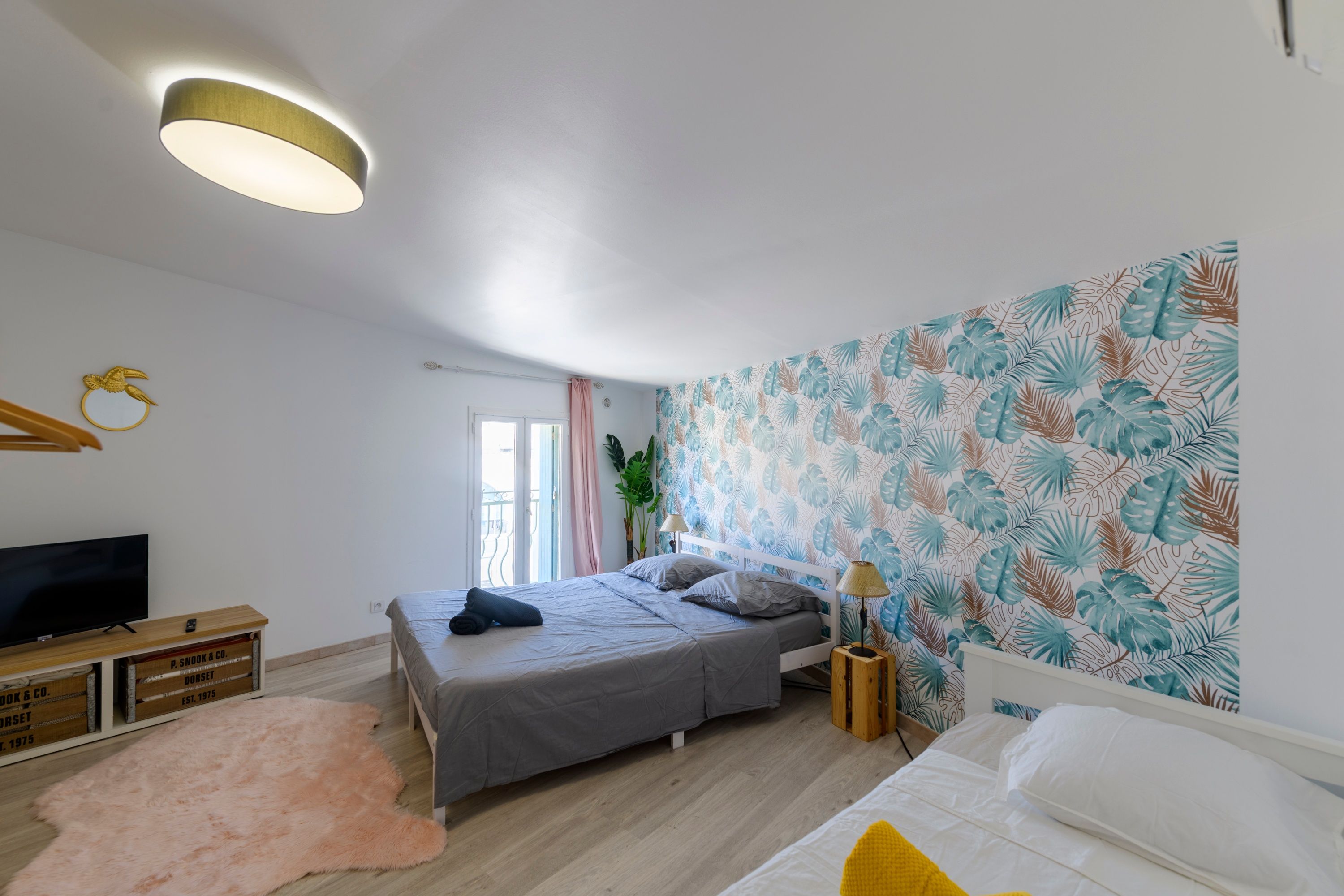 Ein Schlafzimmer mit einem Bett, einem Fernseher, einer Holzkommode, einem rosa Teppich und einer Wand mit tropischen Blättermotiven.
