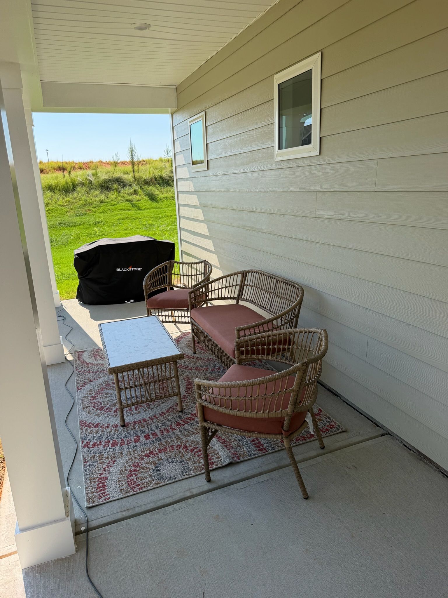 Un patio couvert avec une table et quatre chaises en osier, un barbecue et une vue sur un champ verdoyant.