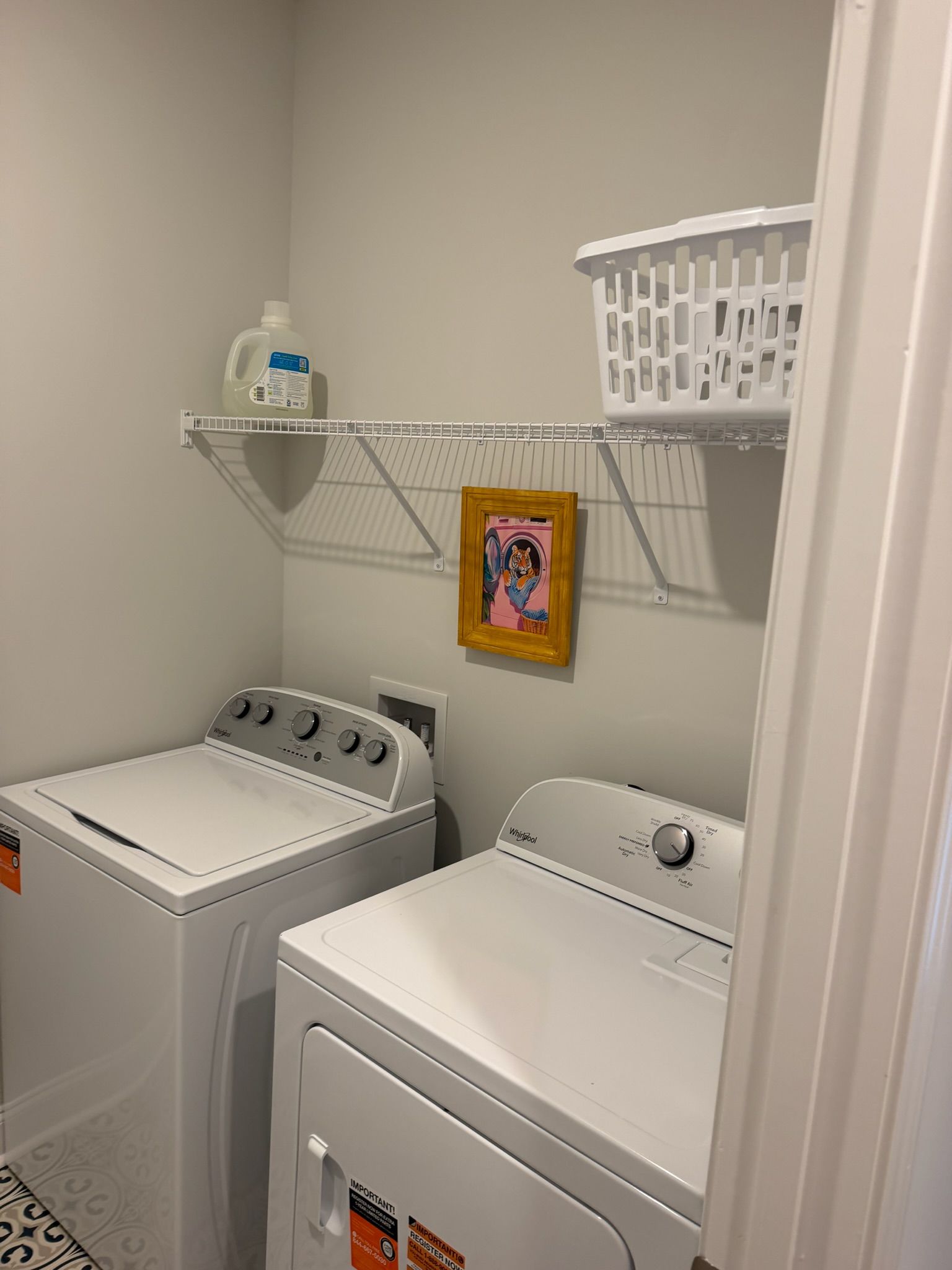 Uma lavanderia com duas máquinas de lavar e secar de abertura frontal, uma prateleira com uma cesta e uma garrafa, e um quadro na parede.