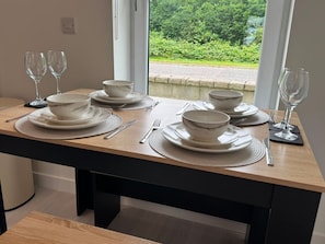 A dining table set with white plates, bowls, cutlery, and wine glasses, positioned near a window with a view of greenery. - Duthie Gardens Retreat – Peterhead (Aberdeenshire)
