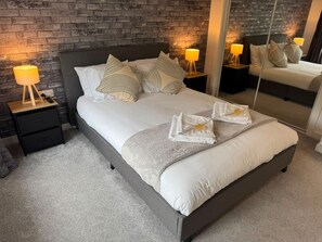A modern bedroom with a large bed, two bedside tables with lamps, a mirror, and a brick wall. - Duthie Gardens Retreat – Peterhead (Aberdeenshire)