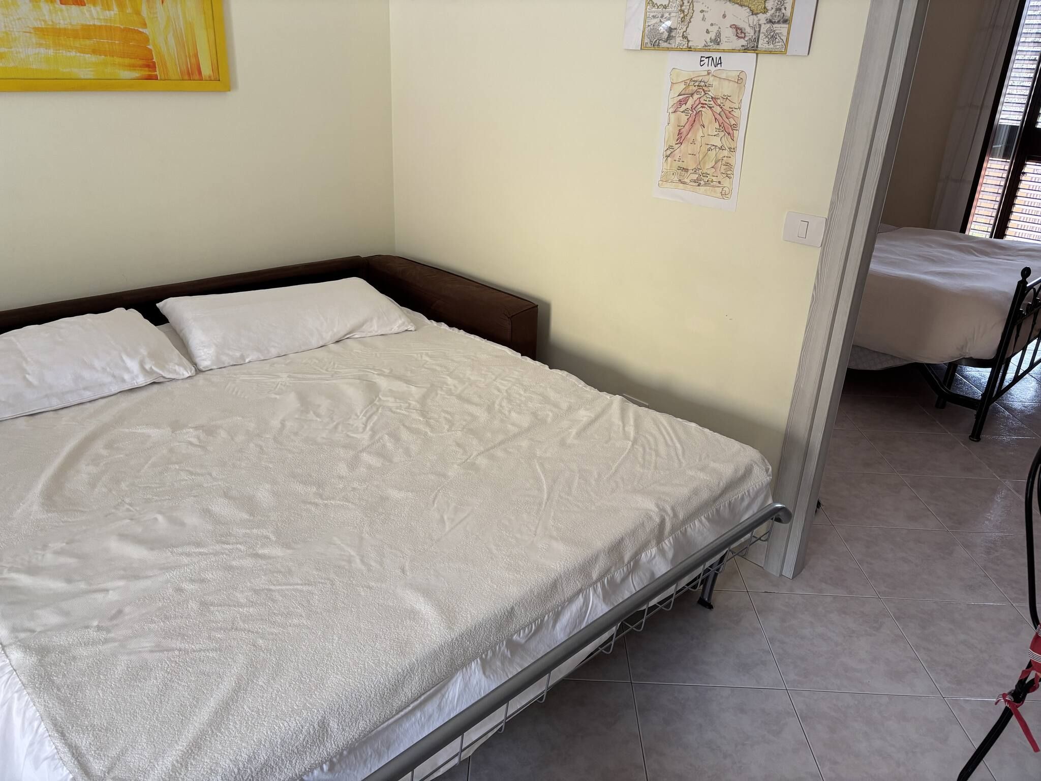 Una cama individual con estructura metálica, colchón blanco y cabecera. Se aprecia un mapa colgado en la pared y un interruptor de luz.