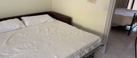 Una cama individual con estructura metálica, colchón blanco y cabecera. Se aprecia un mapa colgado en la pared y un interruptor de luz.