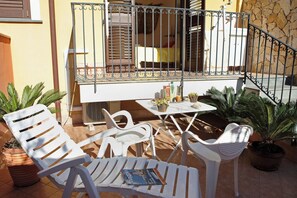 Varanda com cadeiras de plástico branco, uma mesa e um pequeno vaso de planta, além de corrimão de metal.