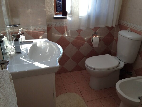 Casa de banho com lavatório branco, sanita e bidé. Tem uma janela com cortinas e o chão tem azulejos estampados.