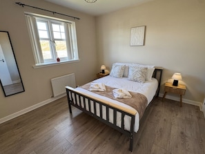 A bedroom with a wooden bed, two bedside tables with lamps, a window with curtains, and a radiator.