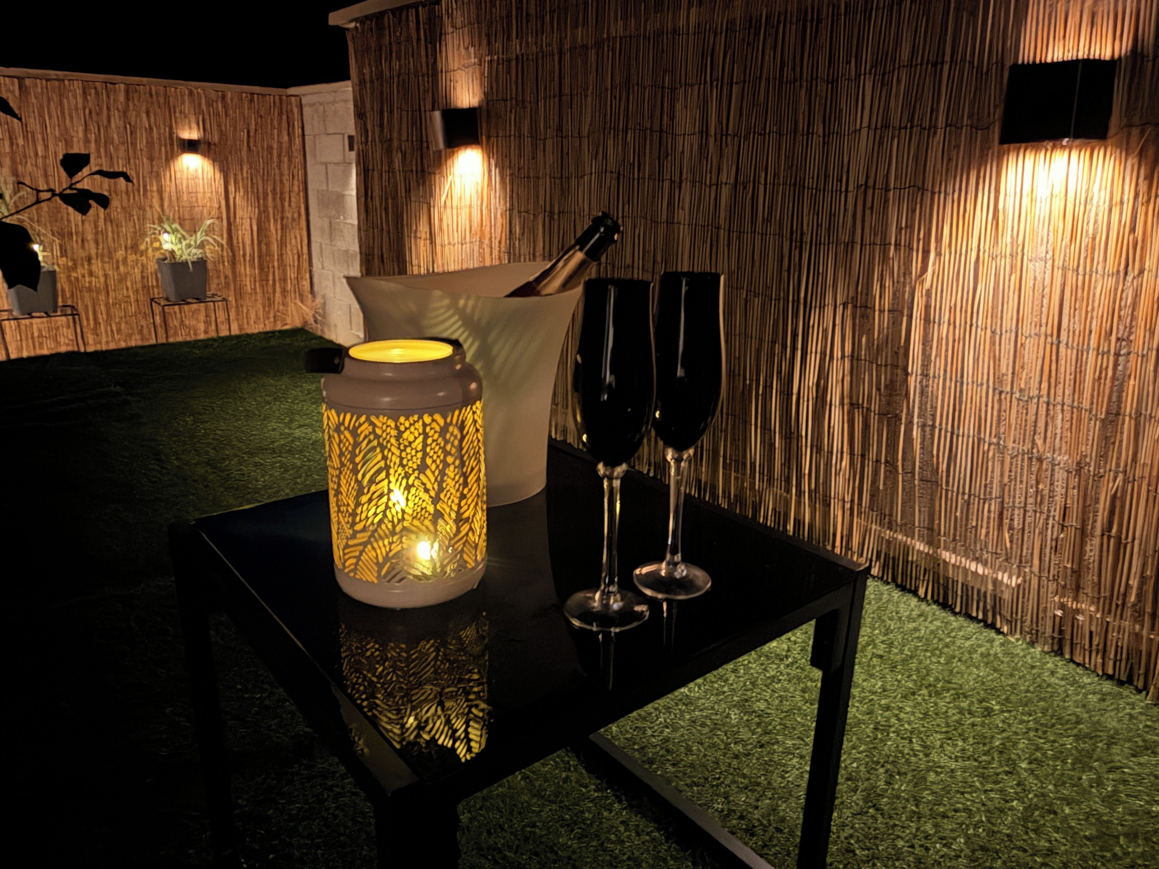 Une table ornée d’une lanterne décorative, de deux verres à vin et d’un chandelier, dans une pièce dont le mur est recouvert de panneaux de bambou et le sol, de gazon synthétique.
