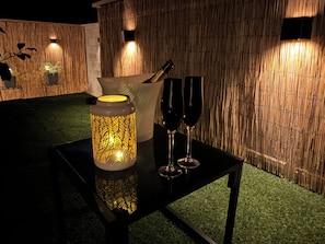 A table with a decorative lantern, two wine glasses, and a candle holder in a room with a bamboo wall and artificial grass flooring. - Le Bain des Violettes • Suite romantique avec jacuzzi privatif (Dole)