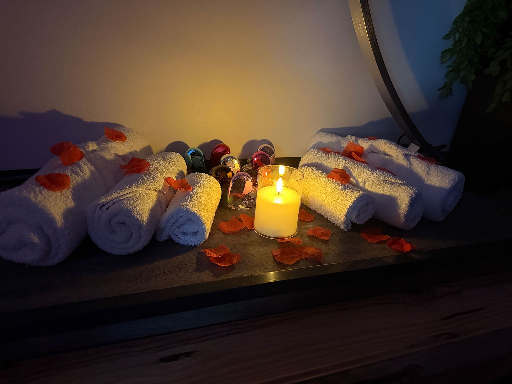 A wooden surface with neatly rolled white towels, a lit candle, and scattered rose petals.