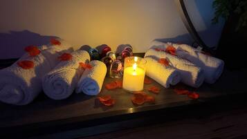 A wooden surface with neatly rolled white towels, a lit candle, and scattered rose petals.