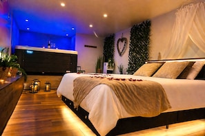 A modern hotel room with a large bed, a minibar, a wall-mounted TV, and a decorative plant. - Le Bain des Violettes • Suite romantique avec jacuzzi privatif (Dole)