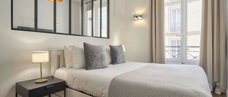Une chambre d’hôtel avec un grand lit, une table de chevet avec une lampe, un miroir et une vue sur la ville par la fenêtre.