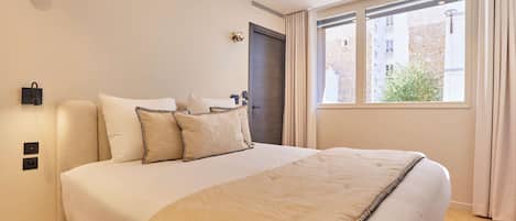 A hotel room with a bed, a window with curtains, a door, and a wall-mounted light fixture.