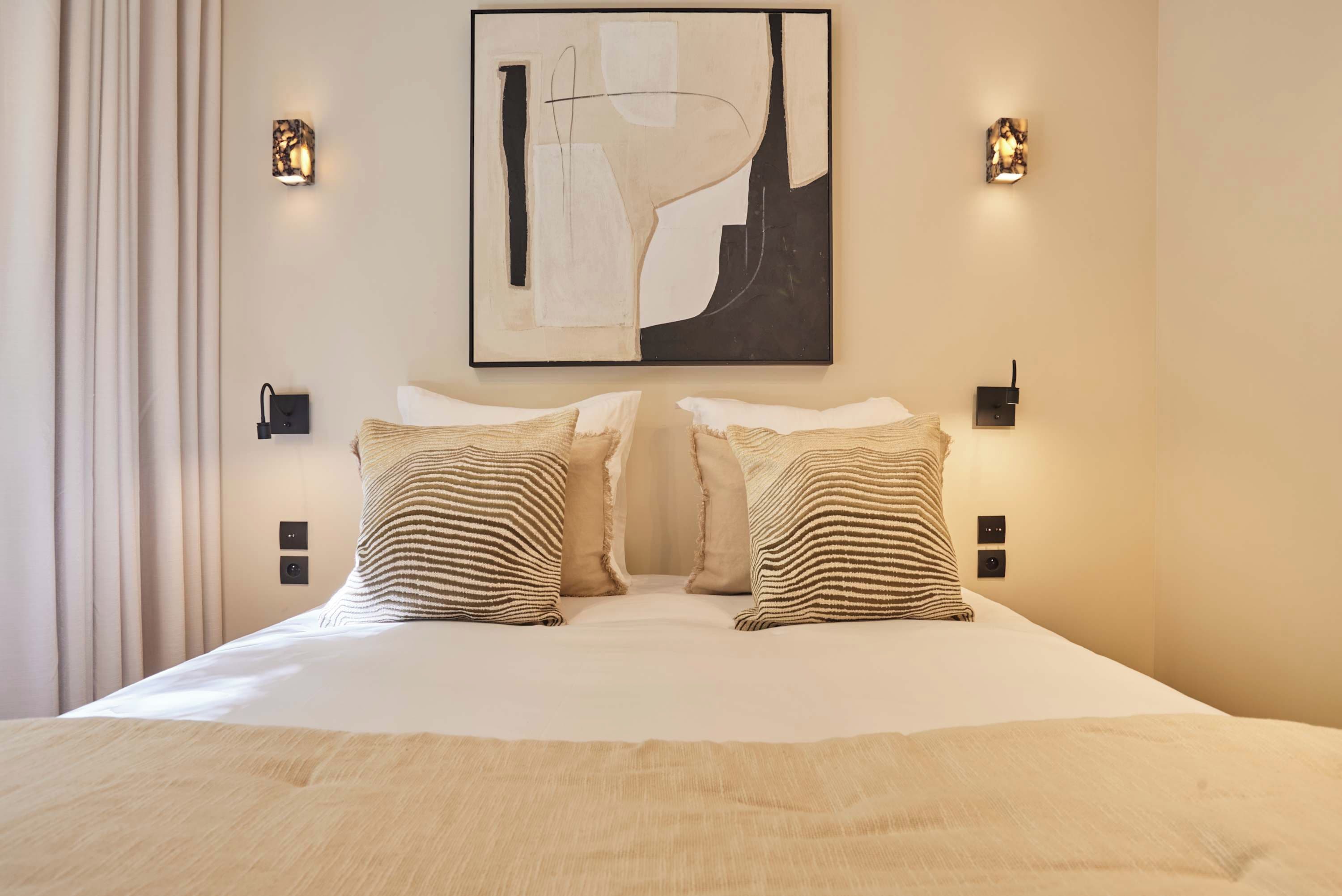 Une chambre d’hôtel avec un grand lit, deux oreillers, un tableau abstrait accroché au mur et deux appliques murales.