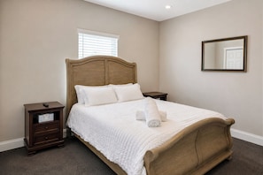 A neatly made bed with white bedding and a wooden headboard in a room with beige walls, a window with blinds, and a mirror on the wall. - Your Tampa Home Away From Home. 3BR/2BA (Tampa)