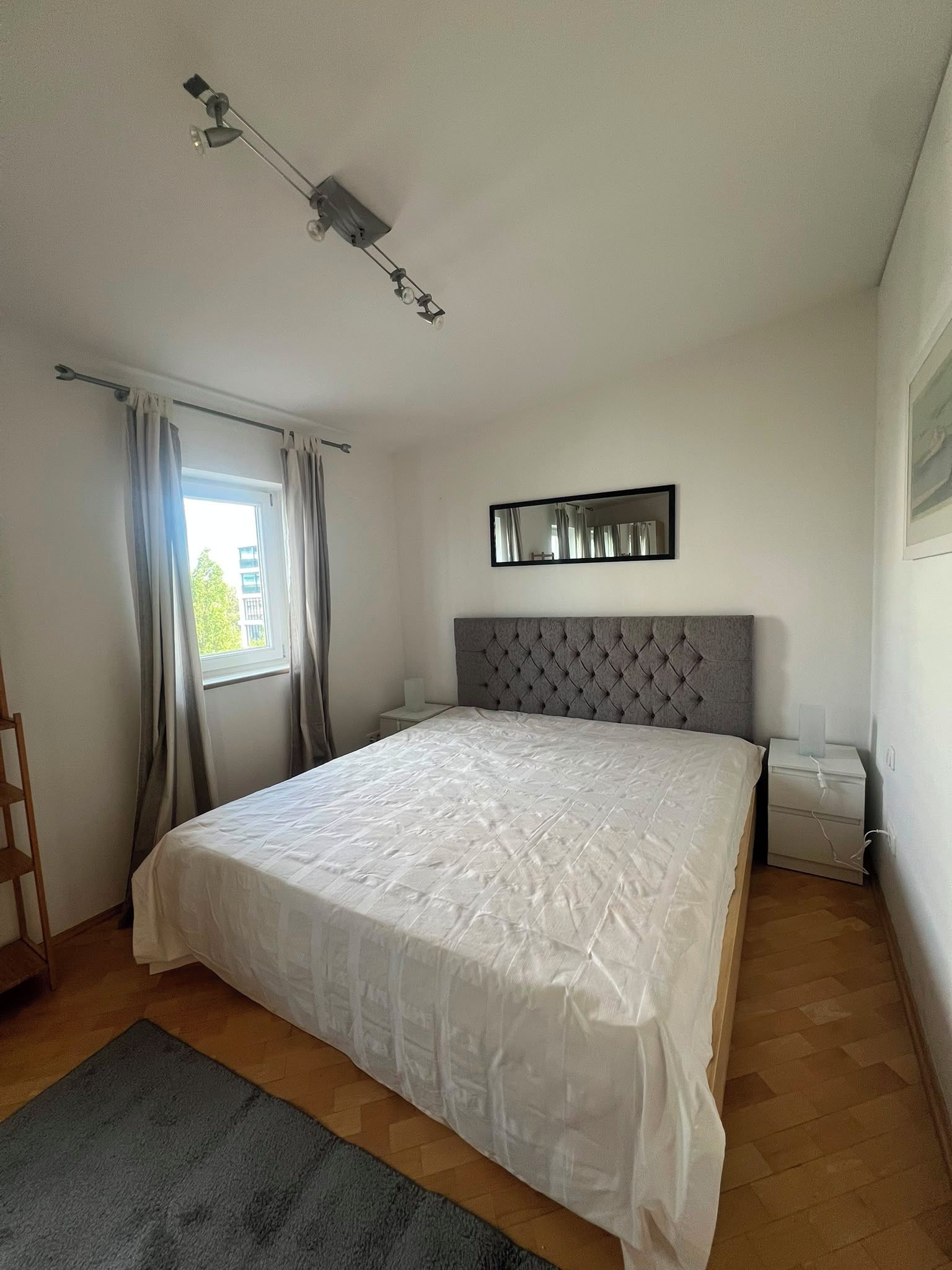 Ein Schlafzimmer mit einem großen Bett, einem Fenster mit Vorhängen, einem Nachttisch, einem wandmontierten Spiegel und einer Deckenleuchte.