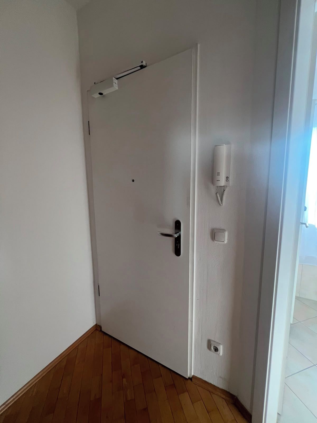Eine weiße, geschlossene Tür mit einem Metallgriff, einem weißen Gegensprechanlage und einem Lichtschalter an der Wand.