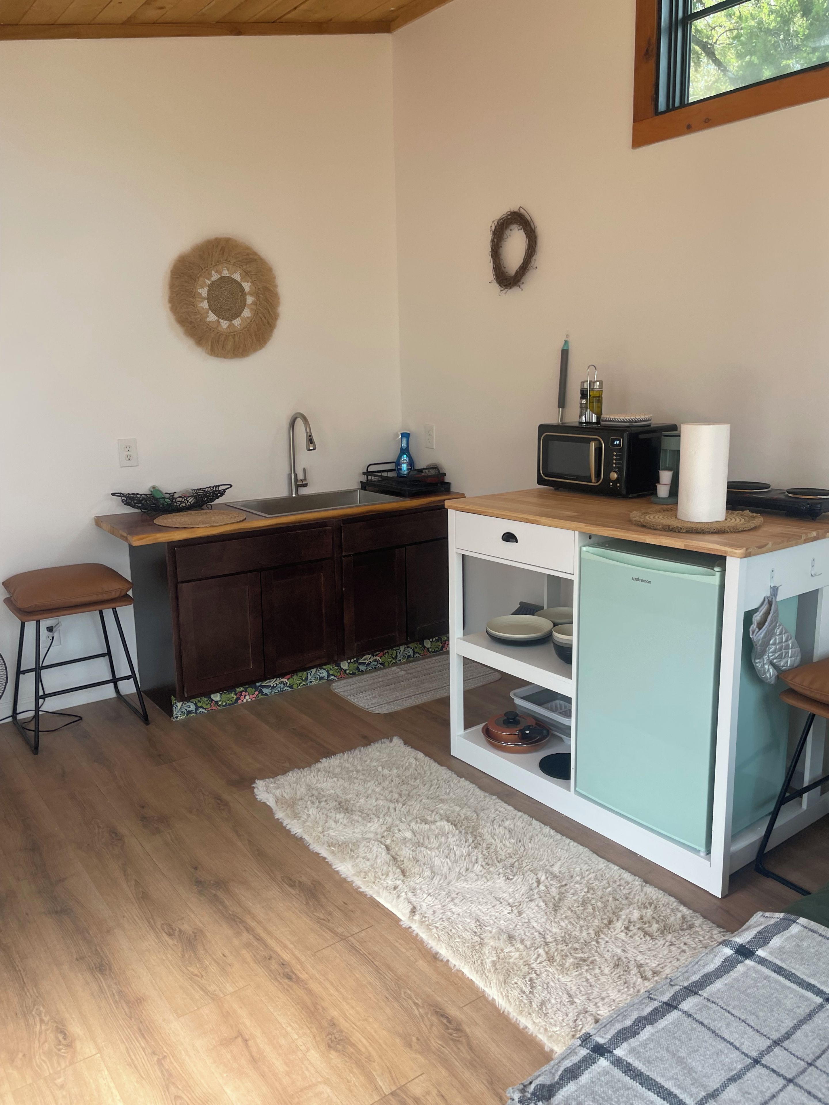 A compact kitchen with a sink, microwave, and a small refrigerator. There are stools and a rug on the floor.