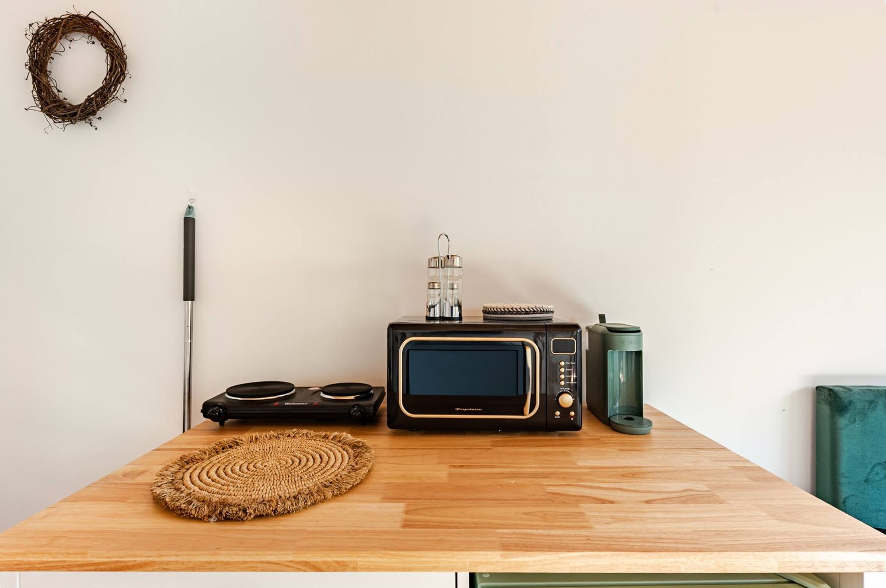 A wooden table with a woven placemat, a black microwave, a green mug, and a black speaker.