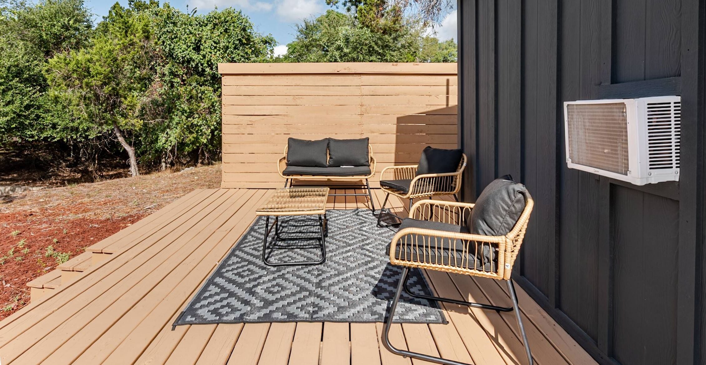 An outdoor patio with a wooden deck, a sofa, two chairs, and a patterned rug.