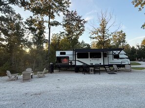 A Kodiak Ultimate RV parked on gravel with outdoor seating and a covered area. - Adventure Awaits! Come Camp, Glamp, and RV with us! Golf cart included! (Corbin)