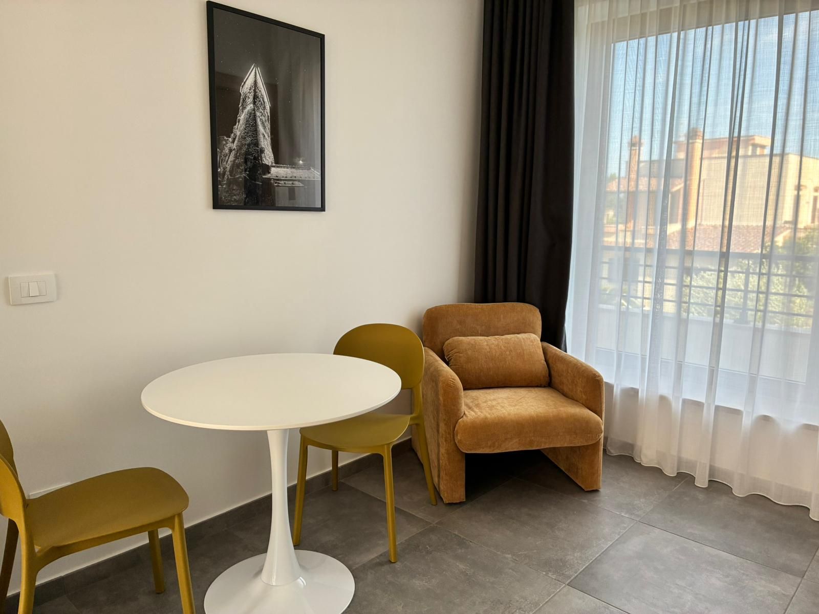 Une pièce avec une table ronde, deux chaises, un canapé, une fenêtre avec des rideaux légers et un tableau encadré au mur.