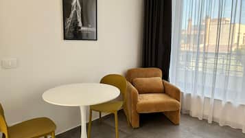Une pièce avec une table ronde, deux chaises, un canapé, une fenêtre avec des rideaux légers et un tableau encadré au mur.