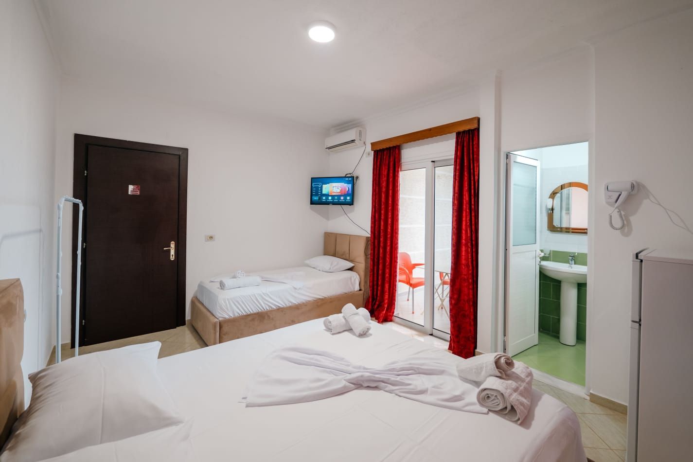 A hotel room with two beds, a TV, a sliding glass door to a balcony, and a bathroom with a sink and mirror.