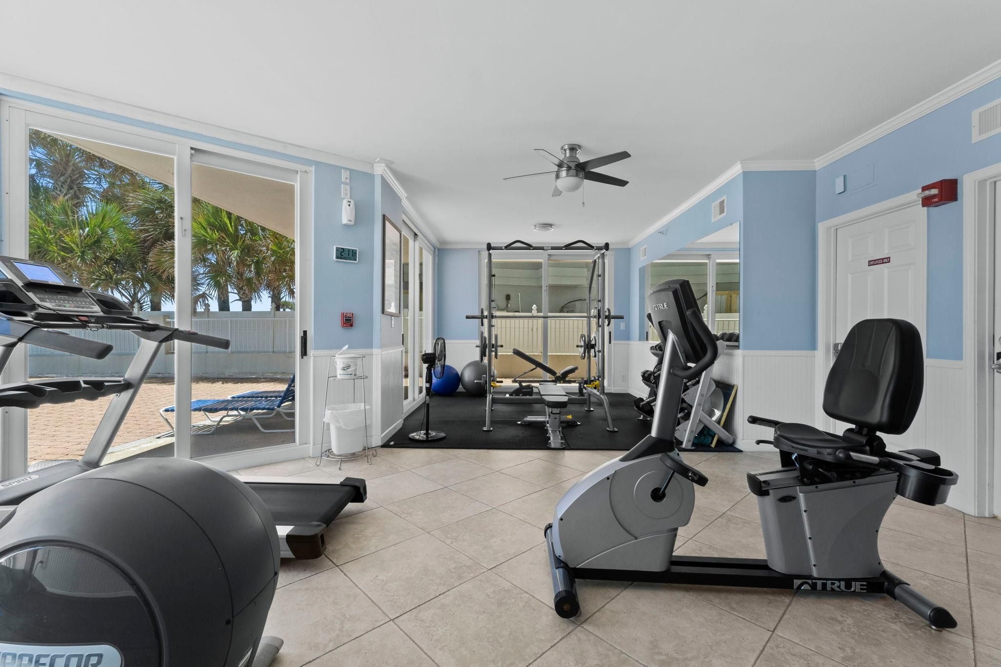 Ein Fitnessraum mit Trainingsgeräten, einem Deckenventilator und Blick auf den Strand durch große Fenster.