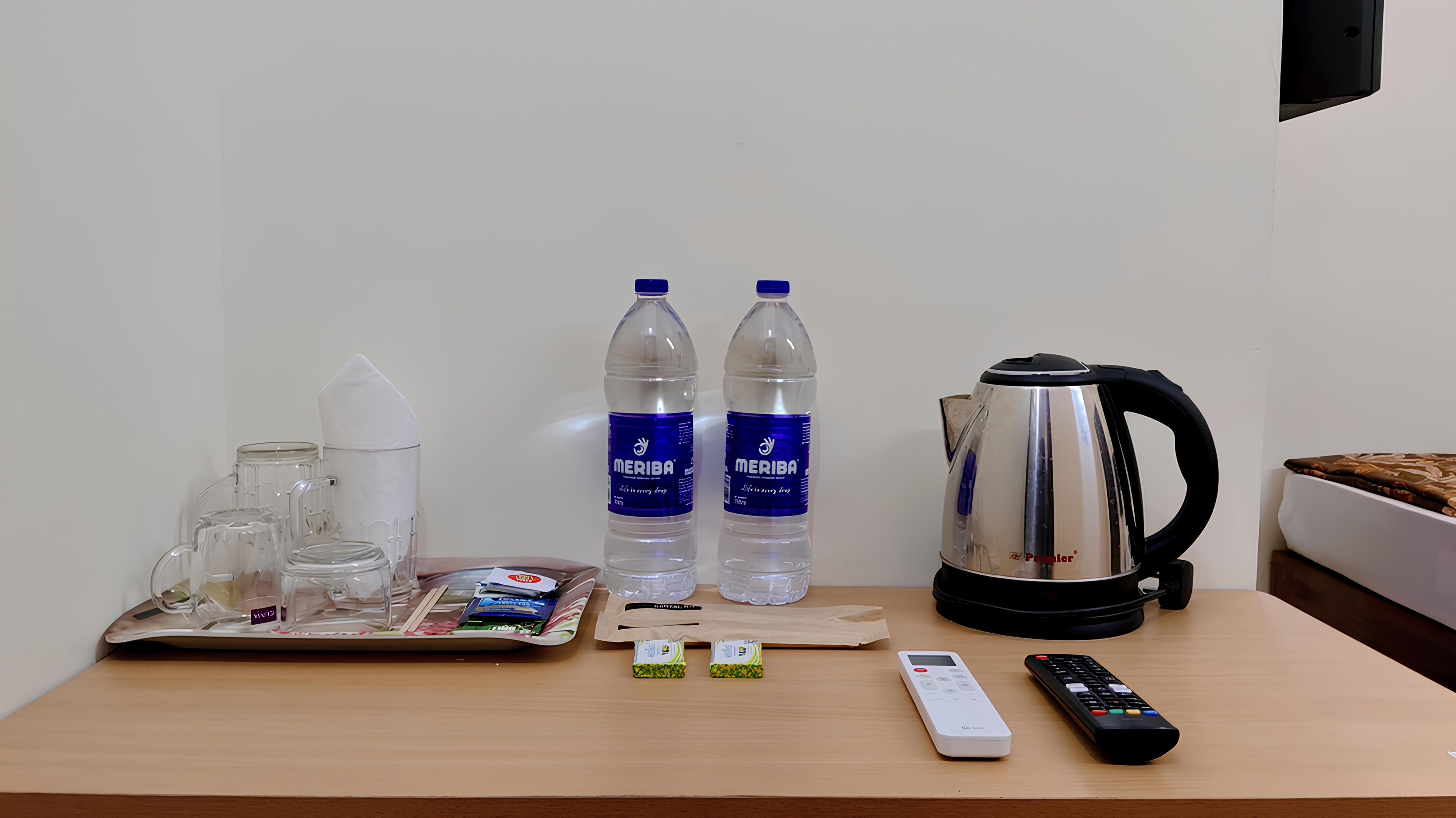 Habitación de hotel con una mesa de madera, dos botellas de agua, una tetera, una bandeja con vasos y pañuelos de papel, un control remoto y un pequeño dispositivo electrónico.