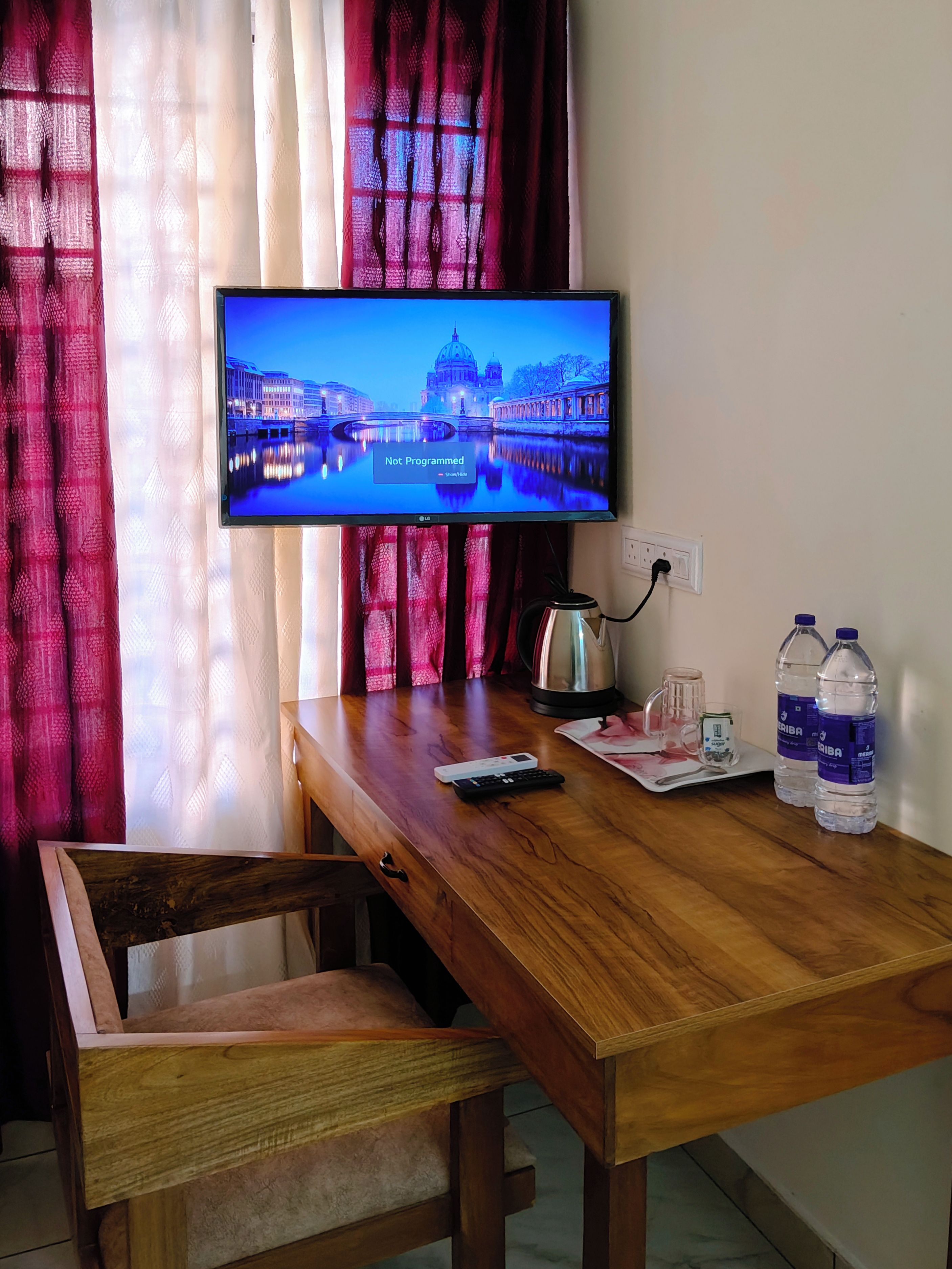 A room with a wooden desk, a television displaying a cityscape, a kettle, and bottled water.