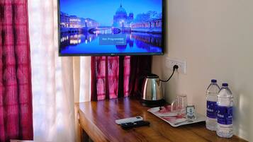 A room with a wooden desk, a television displaying a cityscape, a kettle, and bottled water.
