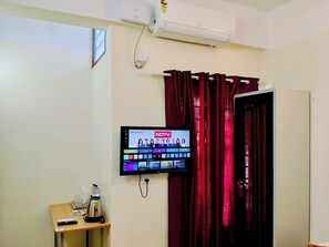 A hotel room with a flat-screen TV mounted on the wall, a small table with a kettle, and a red curtain.