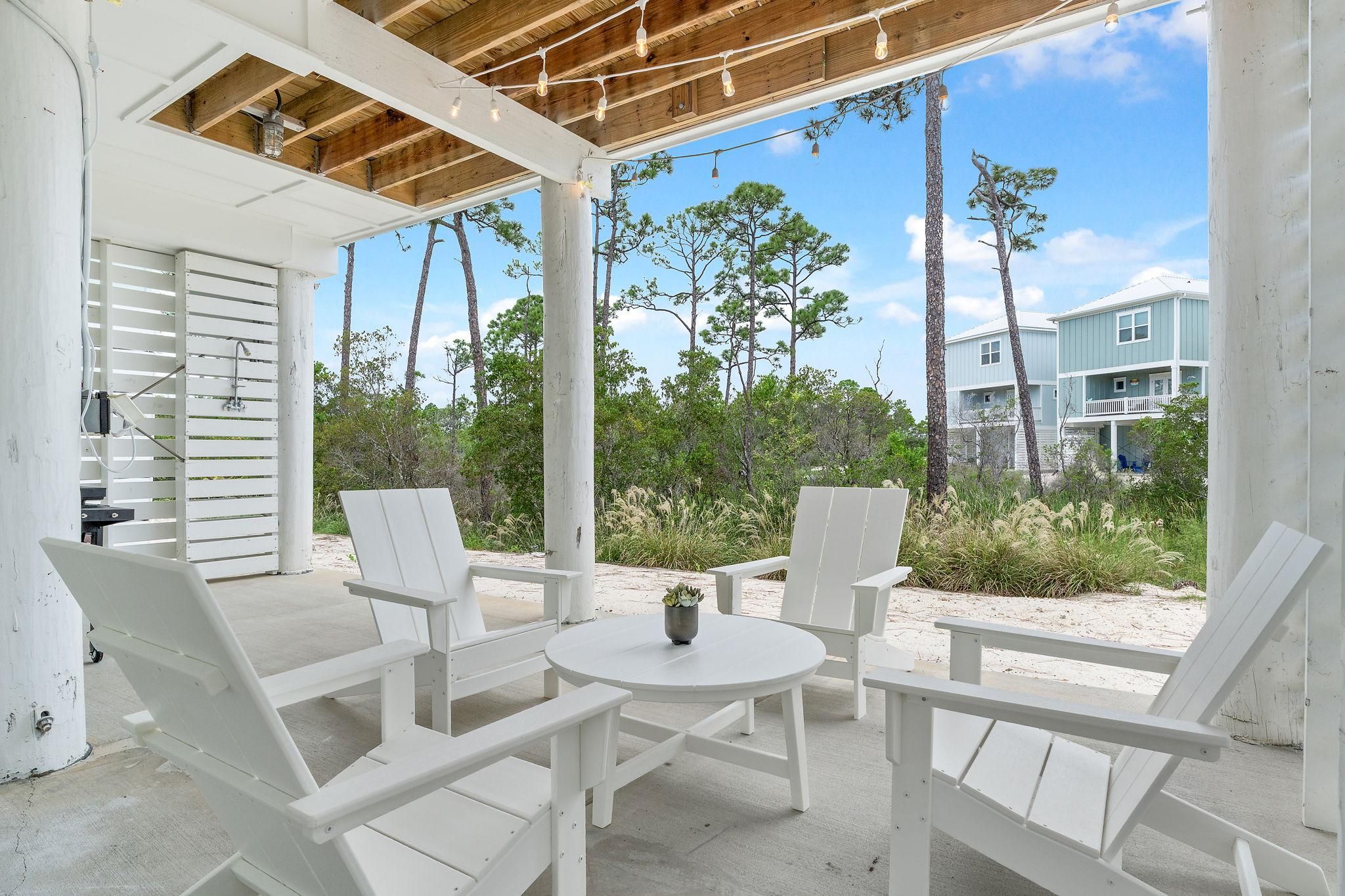 A covered outdoor seating area with white chairs and a table, overlooking a beach with trees and a building in the background.
